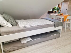 zaścielone łóżko, pod nim dodatkowy materac, w rogu stół z krzesłami