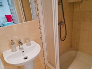 łazienka z prysznicem, w lustrze odbija się łóżko z czerwoną poduszką